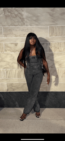 a woman wearing overalls and heels stands in front of a brick wall