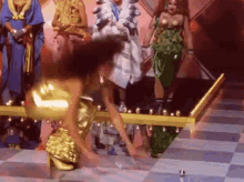 a woman in a gold dress is standing on a checkered floor in front of a group of women in costumes .