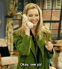 a woman in a green coat is smiling while talking on a phone and says okay see ya