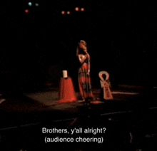 a woman singing into a microphone on a stage with the words brothers y all alright ( audience cheering ) below her