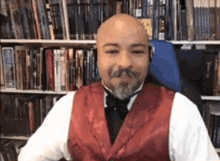 a bald man with a beard wearing a red vest and bow tie is sitting in front of a bookshelf .