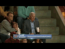 a man in a suit sits in a stadium watching a football game with the name frank beamer above him