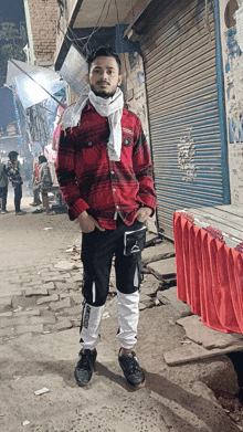 a young man wearing a red plaid shirt and black pants is standing on a cobblestone street with his hands in his pockets