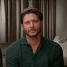 a man with a beard wearing a green polo shirt is sitting on a couch .