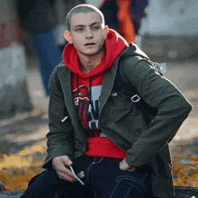 a young man wearing a red hoodie and a green jacket is sitting on the ground smoking a cigarette .