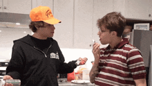 two men in a kitchen one wearing an orange hat that says ' fgm ' on it