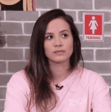 a woman in a pink shirt is sitting in front of a brick wall with a sign that says feminino .