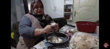 a woman in a hijab is preparing food in a frying pan on a table next to a bag of noodles