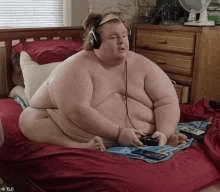 a shirtless fat man is sitting on a bed playing a video game .