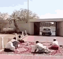 a group of men are sitting on a rug in front of a building with a car driving by .