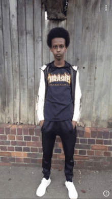 a young man wearing a thrasher shirt stands in front of a wooden fence