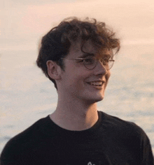 a young man with curly hair and glasses is smiling while standing in front of the ocean .