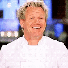 a man in a white chef 's coat is smiling in front of a blurry background .