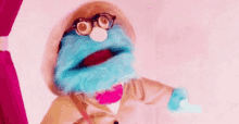 a blue puppet with glasses and a hat is standing in front of a pink curtain and looking at the camera .