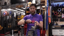 a man in a purple shirt is working on a piece of metal on a vise in a garage .