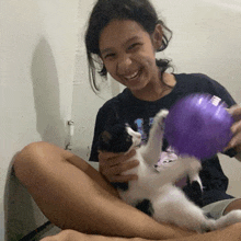 a girl in a black shirt is holding a cat and a purple ball