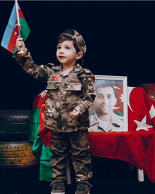 a little boy in a military uniform is holding a flag in front of a picture of a soldier