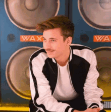 a young man with a mustache is sitting in front of a wall of speakers with the word wax on it .