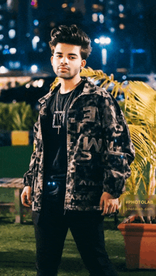 a man wearing a black shirt and a camo jacket stands in front of a palm tree