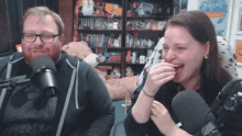 a man and a woman are laughing in front of a microphone while sitting in front of a bookshelf with stuffed animals