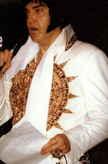 a man in a white and gold suit holds a microphone and a piece of paper