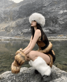 a woman wearing a fur hat and leg warmers is squatting on a rock near a lake