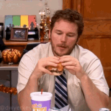 a man sitting at a table eating a hamburger and drinking a purple cup that says ' king ' on it