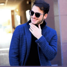 a man wearing sunglasses and a blue jacket is smiling and touching his chin .