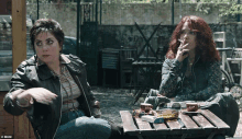 two women are sitting at a table smoking cigarettes and drinking coffee