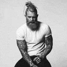 a man with a beard and tattooed arms is wearing a white t-shirt and a top knot .