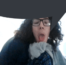 a person with curly hair wearing glasses and a plaid shirt sticks out their tongue