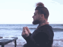 a man with a tattoo on his neck sits in front of the ocean with his hands folded in prayer