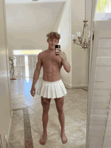 a shirtless man in a white pleated skirt takes a picture of himself in the mirror