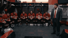 a group of hockey players are sitting in a locker room with a man in a suit talking to them