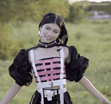 a woman wearing a choker and a crop top is standing in a field with her arms outstretched and smiling .