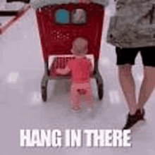 a baby is standing next to a shopping cart in a store and a woman is walking behind it .