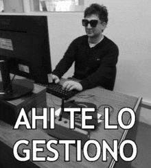 a man wearing sunglasses sits at a desk using a computer with the words " ahi te lo gestiono " below him