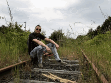 a man sitting on train tracks wearing sunglasses and a black shirt that says ' i love you ' on it