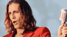 a man with long hair is singing into a microphone while wearing a red jacket