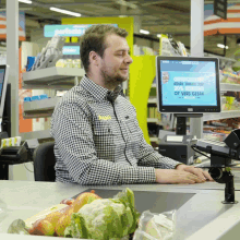 a man in a checkered shirt sits at a cash register in front of a computer screen that says gefeliciteerd