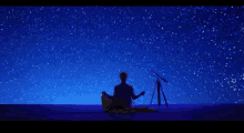 a man sits in a lotus position under a starry night sky next to a telescope