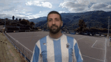 a man wearing a blue and white striped shirt stands in front of a soccer field with mountains in the background