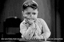 a black and white photo of a little boy sitting at a desk with his hand on his face .