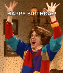 a woman in a colorful sweater is raising her arms in the air and saying `` happy birthday '' .