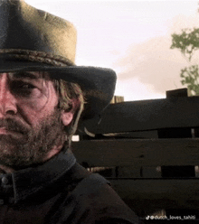 a man with a beard wearing a cowboy hat is standing in front of a wooden fence