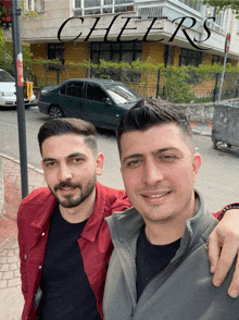 two men are posing for a picture in front of a building that has the word cheers on it