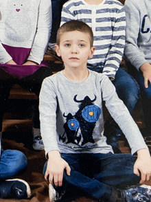 a boy wearing a gray shirt with a bull on it sits on the floor with his legs crossed