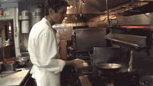 a man in a white coat is cooking in a kitchen with a fire extinguisher on the wall behind him