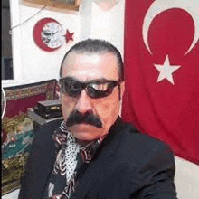 a man with a mustache wearing sunglasses and a suit is taking a selfie in front of a turkey flag .