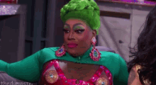 a drag queen with green hair and earrings is sitting next to a woman .